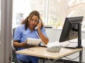 A woman in scrubs, sitting at a desk in a medical exam room, talks on a landline phone.