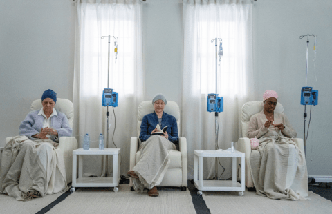 Three seated female patients who are recovering from cancer therapies: one doing her nails, one reading, and the other crocheting.