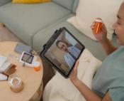 An Asian woman sitting on her couch, a pill bottle in her hand, talking to her doctor whose face can be seen on a tablet computer.